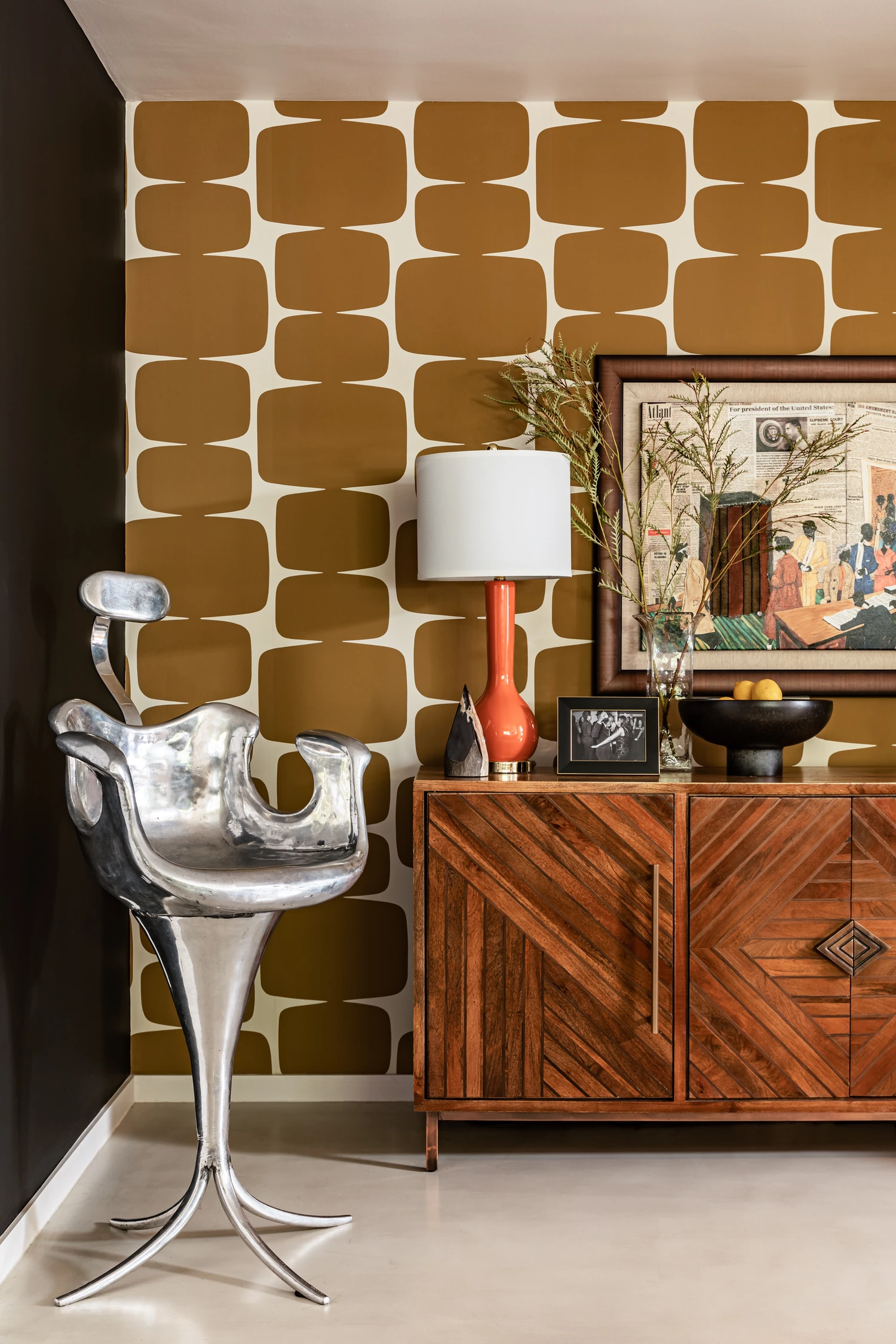 Chrome sculptural chair against gold geometric wallpaper, beside an inlaid credenza