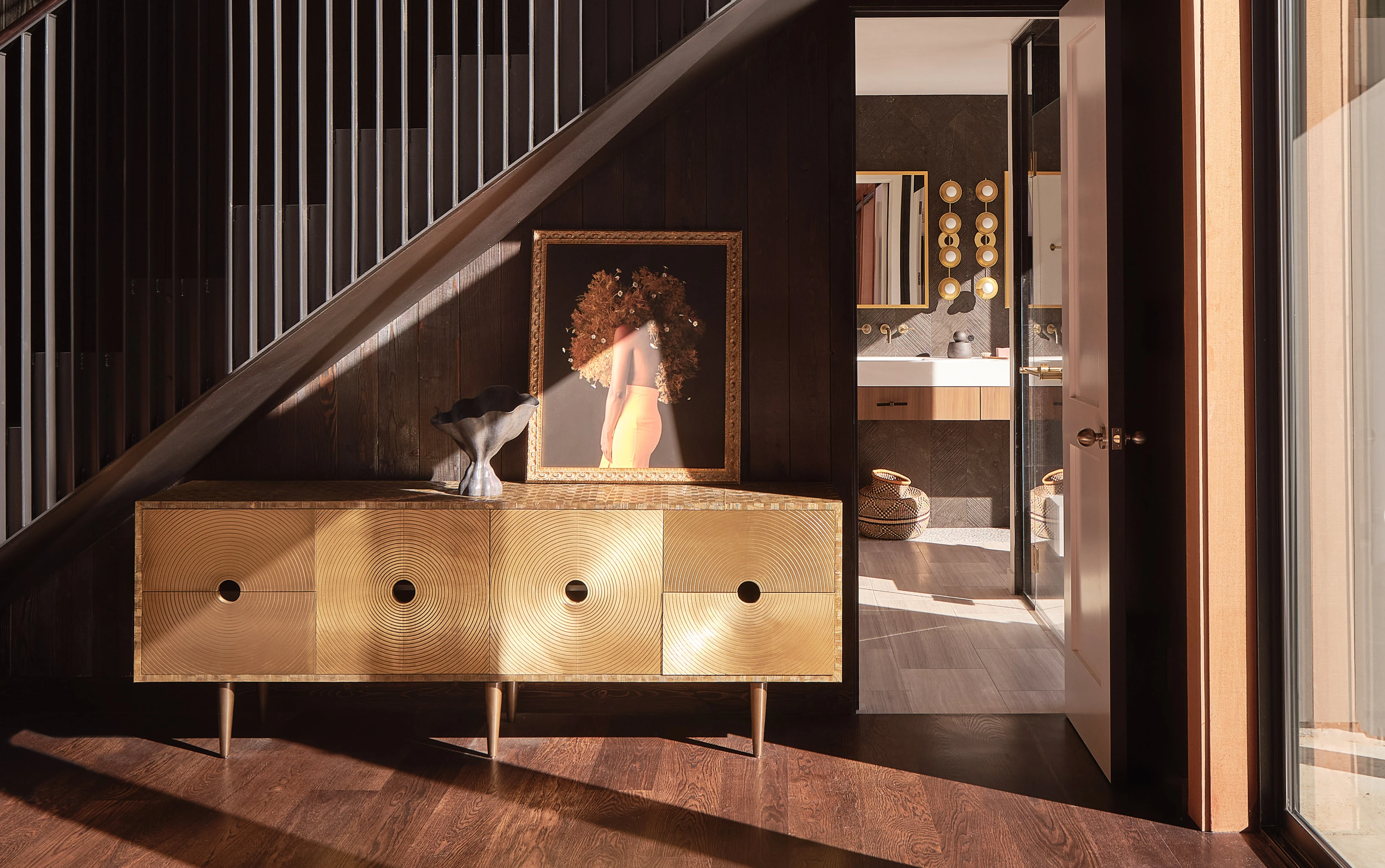 Blackened stair and brass credenza in the Tree House Edge entry, sunlight raking across the floor
